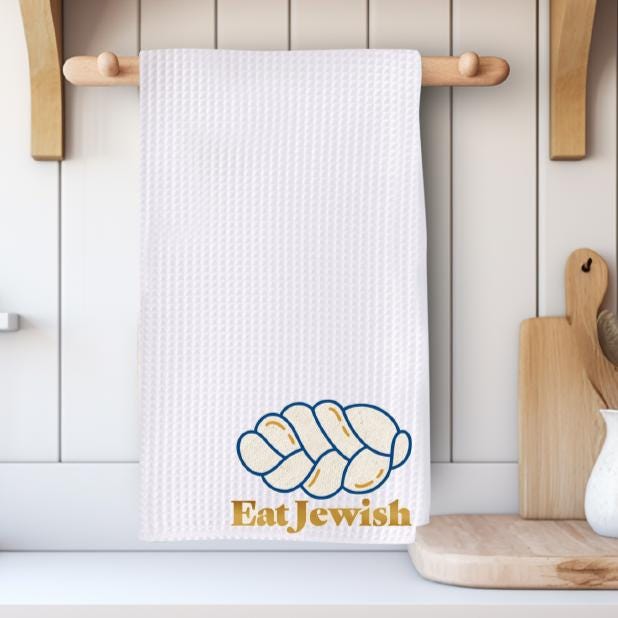 White waffle dish towel with the Eat Jewish logo and challah design in the corner, hanging in a cozy modern kitchen.